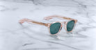 A pair of handcrafted Jacques Marie Mage Zephirin sunglasses in translucent pink with green lenses rest on a white marble surface, showcasing their limited edition appeal.