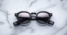 Jacques Marie Mage Zephirin black round sunglasses with thick frames are displayed on a white marble surface.
