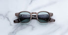 A pair of Jacques Marie Mage Zephirin round sunglasses with dark frames and lenses rests on a light marble surface, showcasing limited edition eyewear by Jacques Marie Mage.