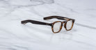 The Jacques Marie Mage Zephirin spectacles by Jacques Marie Mage, featuring handcrafted thick tortoiseshell acetate frames and clear lenses, are elegantly displayed on a white marble surface.