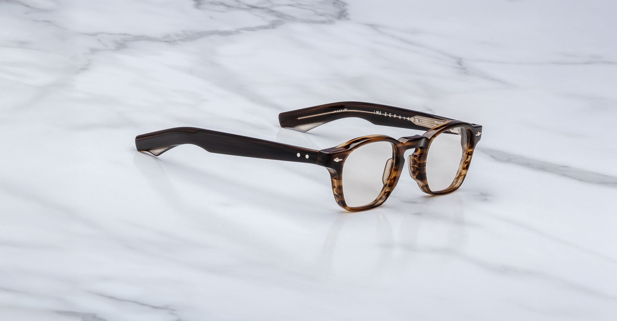 The Jacques Marie Mage Zephirin spectacles by Jacques Marie Mage, featuring handcrafted thick tortoiseshell acetate frames and clear lenses, are elegantly displayed on a white marble surface.