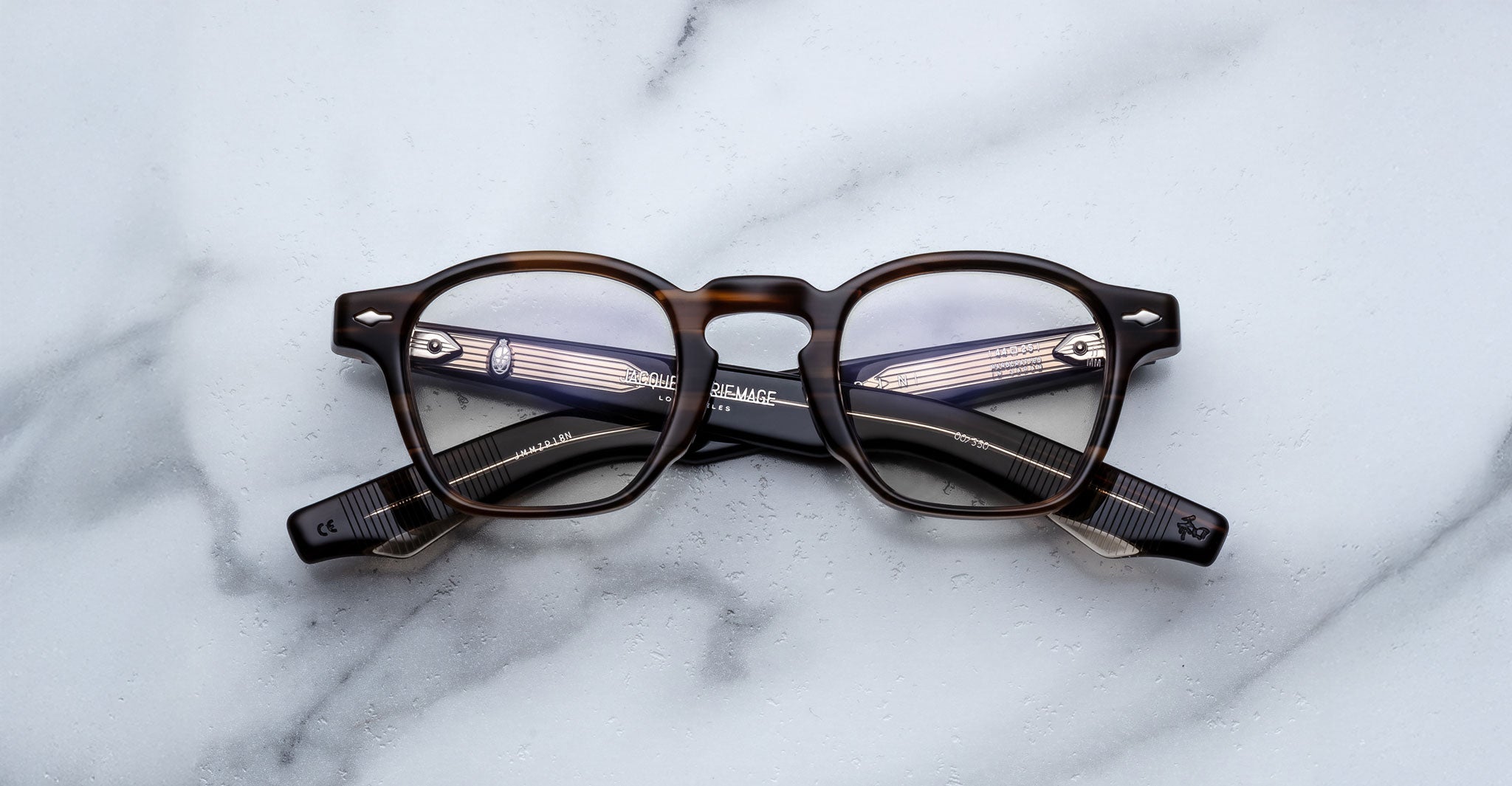 A pair of Jacques Marie Mage Zephirin spectacles with round lenses, handcrafted in acetate, is displayed on a white marble surface.