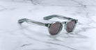 Gray sunglasses with thick translucent frames and dark lenses rest on white marble, evoking the limited edition Jacques Marie Mage Zephirin by Jacques Marie Mage, renowned for distinctive handcrafted acetate eyewear.