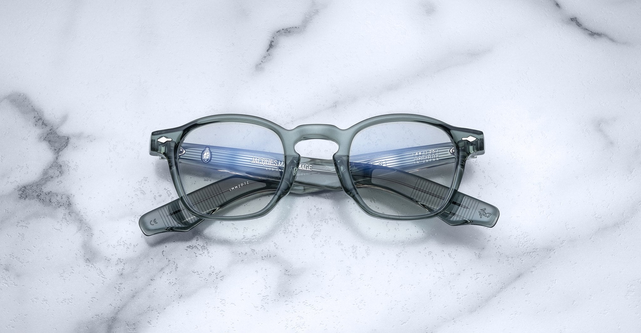 Jacques Marie Mage Zephirin handcrafted translucent gray acetate spectacles with clear lenses rest on white marble, highlighting the brand's refined limited edition eyewear craftsmanship.