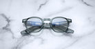 Jacques Marie Mage Zephirin handcrafted translucent gray acetate spectacles with clear lenses rest on white marble, highlighting the brand's refined limited edition eyewear craftsmanship.
