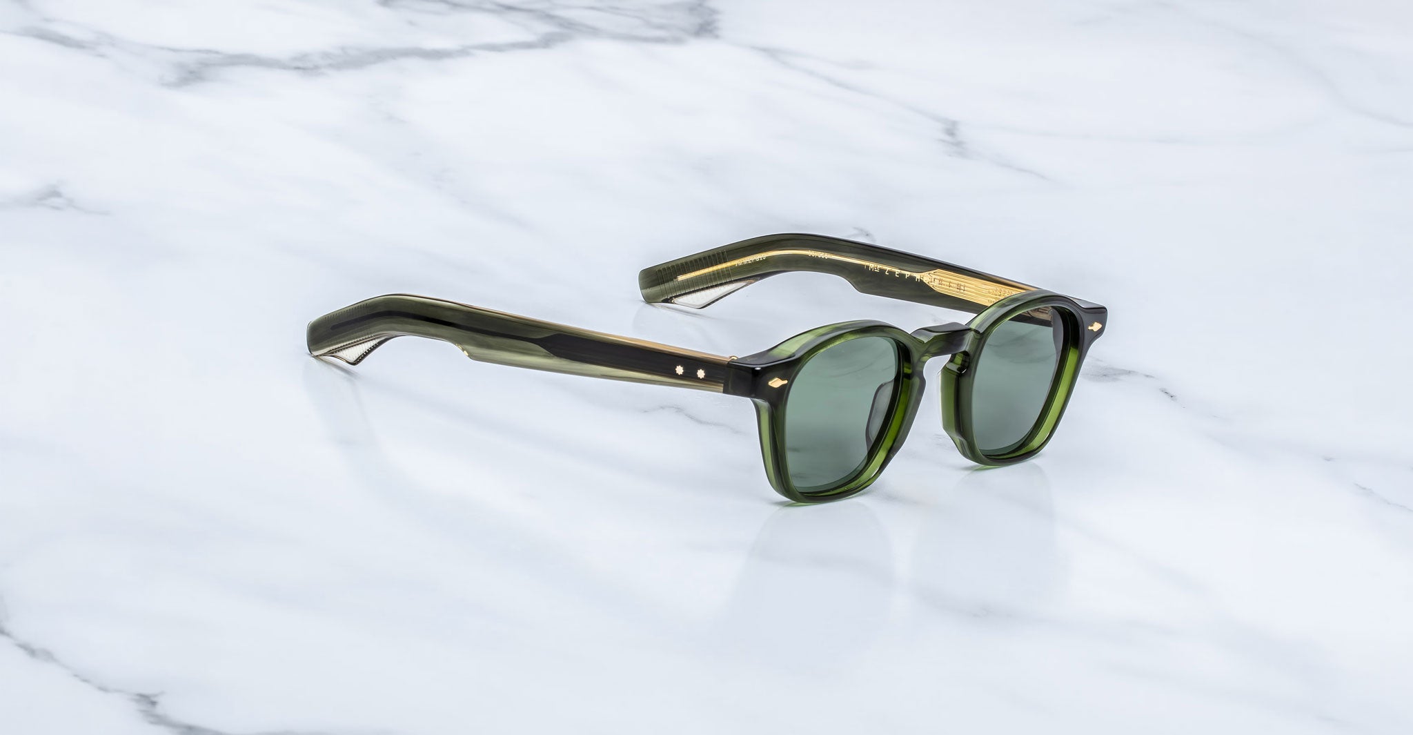 Jacques Marie Mage Zephirin sunglasses in Kombu, three-quarter view with translucent olive green acetate, green-tinted lenses, and gold-tone accents on a white marble surface.