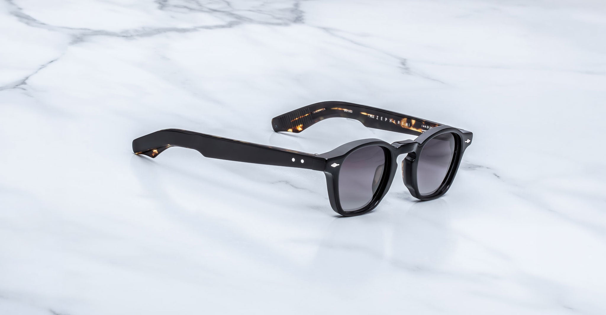 Jacques Marie Mage Zephirin sunglasses in Noir 11, three-quarter view with glossy black acetate, gray-tinted lenses, and subtle tortoiseshell accents on a white marble surface.
