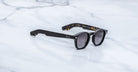 Jacques Marie Mage Zephirin sunglasses in Noir 11, three-quarter view with glossy black acetate, gray-tinted lenses, and subtle tortoiseshell accents on a white marble surface.