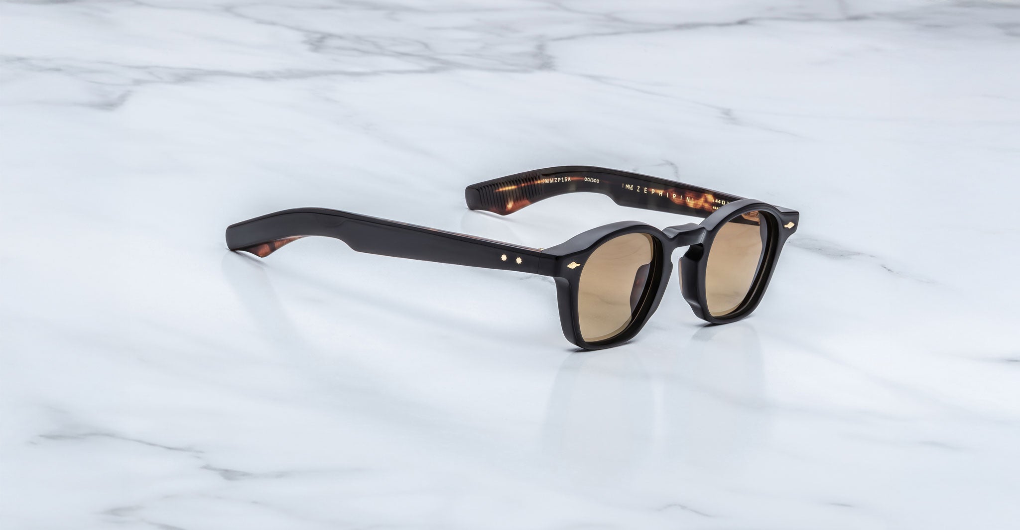 A pair of Jacques Marie Mage Zephirin black sunglasses with brown tinted lenses sits on white marble, highlighting the elegance of limited edition Jacques Marie Mage eyewear.