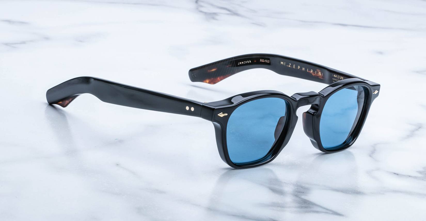 Jacques Marie Mage Zephirin sunglasses feature black acetate, blue lenses, and thick frames, displayed on a white marble surface to highlight the brand’s timeless style in this limited edition eyewear.