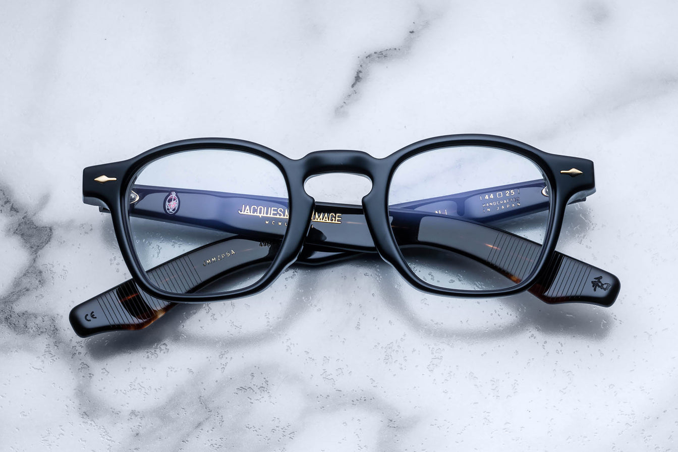 A pair of Jacques Marie Mage Zephirin black rectangular eyeglasses with gold accents, featuring handcrafted acetate frames, are folded and displayed on a white marble surface.