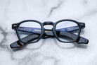 A pair of Jacques Marie Mage Zephirin black rectangular eyeglasses with gold accents, featuring handcrafted acetate frames, are folded and displayed on a white marble surface.