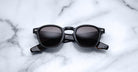 A pair of Jacques Marie Mage Zephirin sunglasses with black square frames and dark lenses sits on white marble, highlighting the brand’s limited edition craftsmanship.