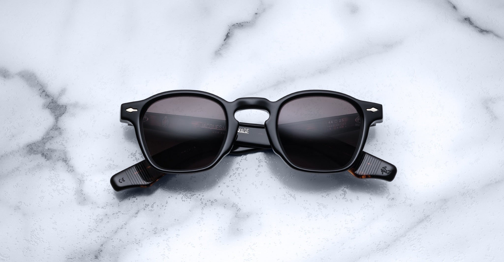 A pair of Jacques Marie Mage Zephirin sunglasses with black square frames and dark lenses sits on white marble, highlighting the brand’s limited edition craftsmanship.