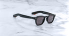 Jacques Marie Mage Zephirin sunglasses feature a classic handcrafted acetate frame with dark lenses, inspired by the brand’s limited edition eyewear, and are displayed on a white marble surface.