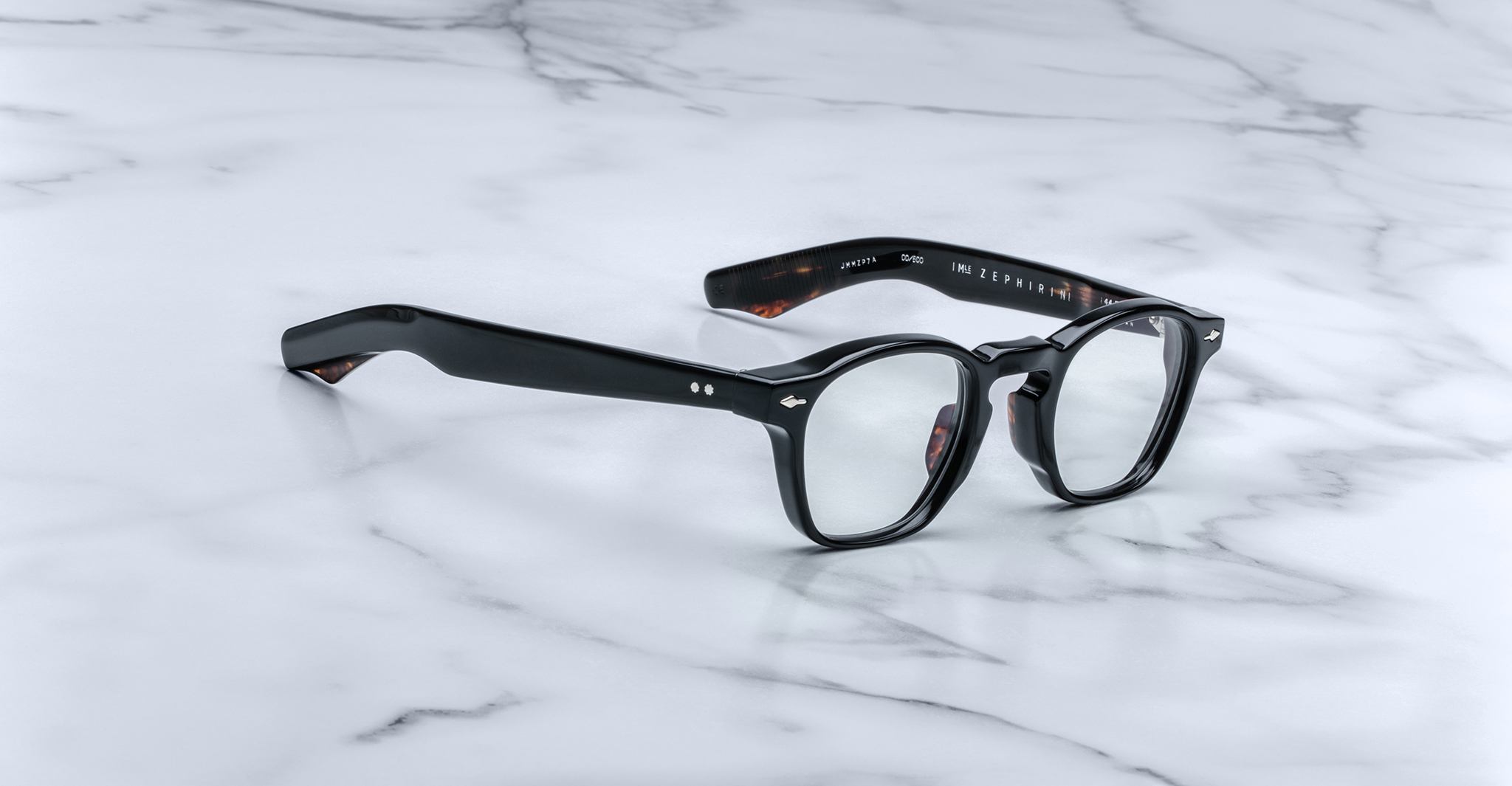 A pair of Jacques Marie Mage Zephirin eyeglasses with black thick rims and clear lenses rests on a white marble surface.