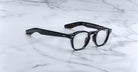 A pair of Jacques Marie Mage Zephirin eyeglasses with black thick rims and clear lenses rests on a white marble surface.