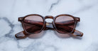 A pair of Jacques Marie Mage Zephirin brown square-framed sunglasses with tinted lenses rests on white marble, highlighting the sophistication of this limited edition eyewear.