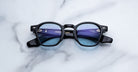 A pair of handcrafted Jacques Marie Mage Zephirin round black eyeglasses with blue-tinted lenses, displayed on a white marble surface.