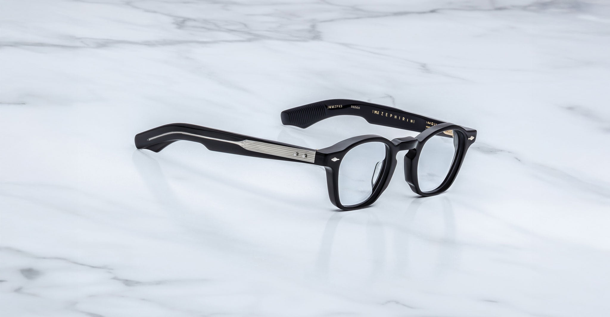 Jacques Marie Mage Zephirin eyeglasses by Jacques Marie Mage, featuring black frames and metal temple detailing, are displayed on white marble, highlighting the handcrafted acetate and striking limited edition design.