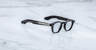 Jacques Marie Mage Zephirin eyeglasses by Jacques Marie Mage, featuring black frames and metal temple detailing, are displayed on white marble, highlighting the handcrafted acetate and striking limited edition design.