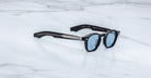 A pair of Jacques Marie Mage Zephirin sunglasses with black frames and blue-tinted lenses is displayed on a white marble surface, highlighting the exclusive style of this limited edition Jacques Marie Mage eyewear.