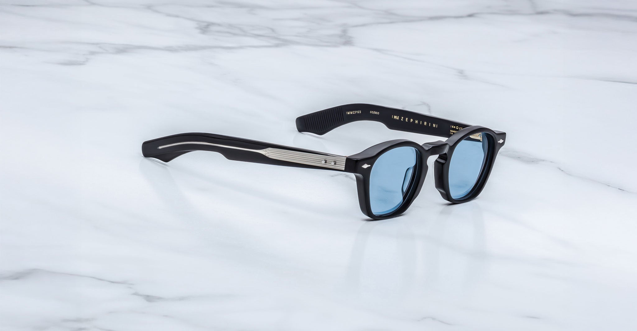 A pair of Jacques Marie Mage Zephirin sunglasses with black frames and blue-tinted lenses is displayed on a white marble surface, highlighting the exclusive style of this limited edition Jacques Marie Mage eyewear.