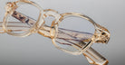 A close-up of Jacques Marie Mage Zephirin spectacles—transparent, beige-tinted handcrafted acetate frames with clear lenses and patterned arms—rests on a reflective surface against a plain background.