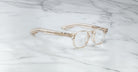 A pair of limited edition Jacques Marie Mage Zephirin eyeglasses in clear, beige-tinted handcrafted acetate with thick frames rests on white marble, showcasing the brand’s signature design and luxury craftsmanship.