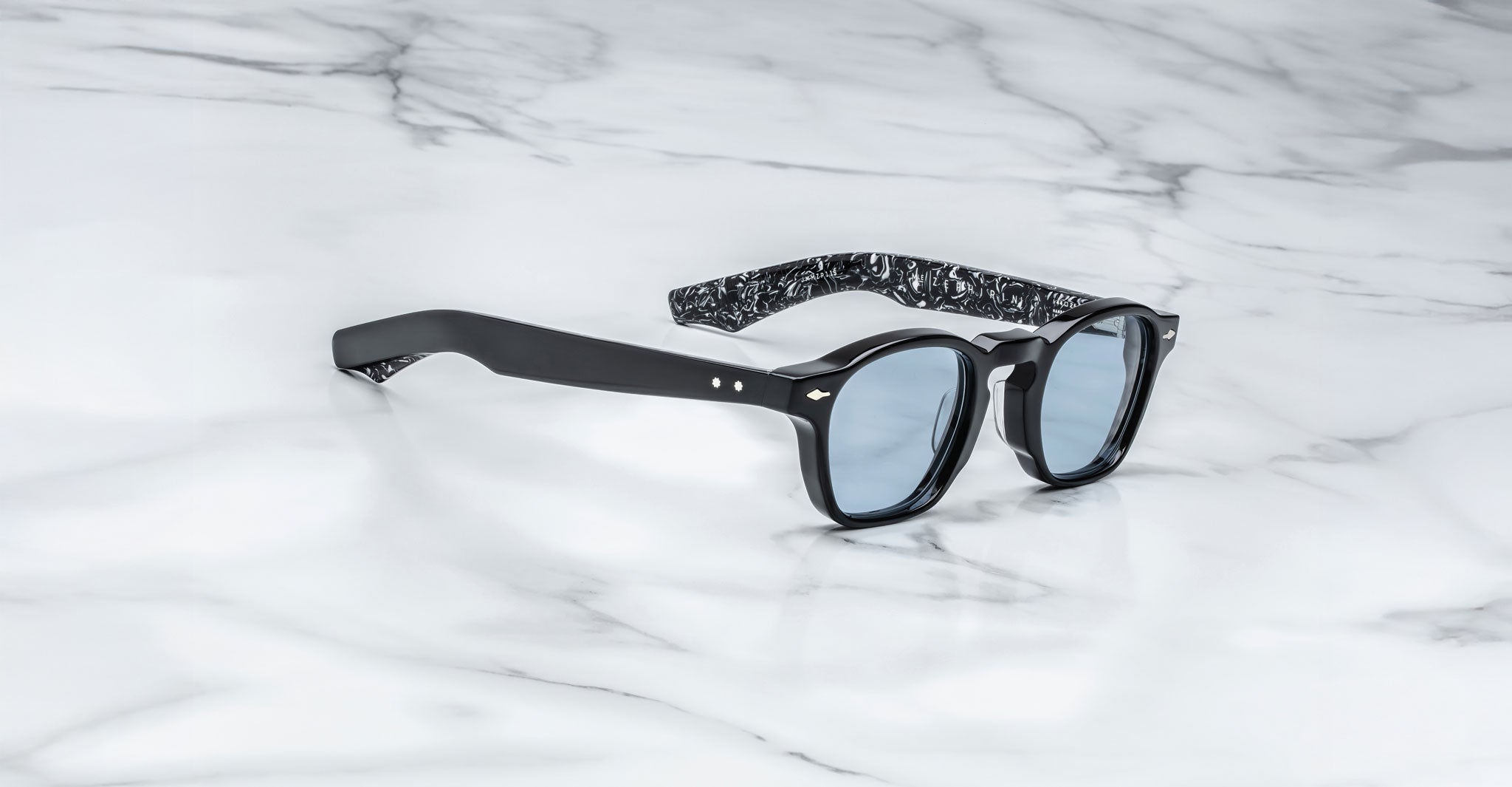 Jacques Marie Mage Zephirin sunglasses feature blue-tinted lenses and marbled inner temples in handcrafted acetate, beautifully displayed on white marble—a striking limited edition by Jacques Marie Mage.