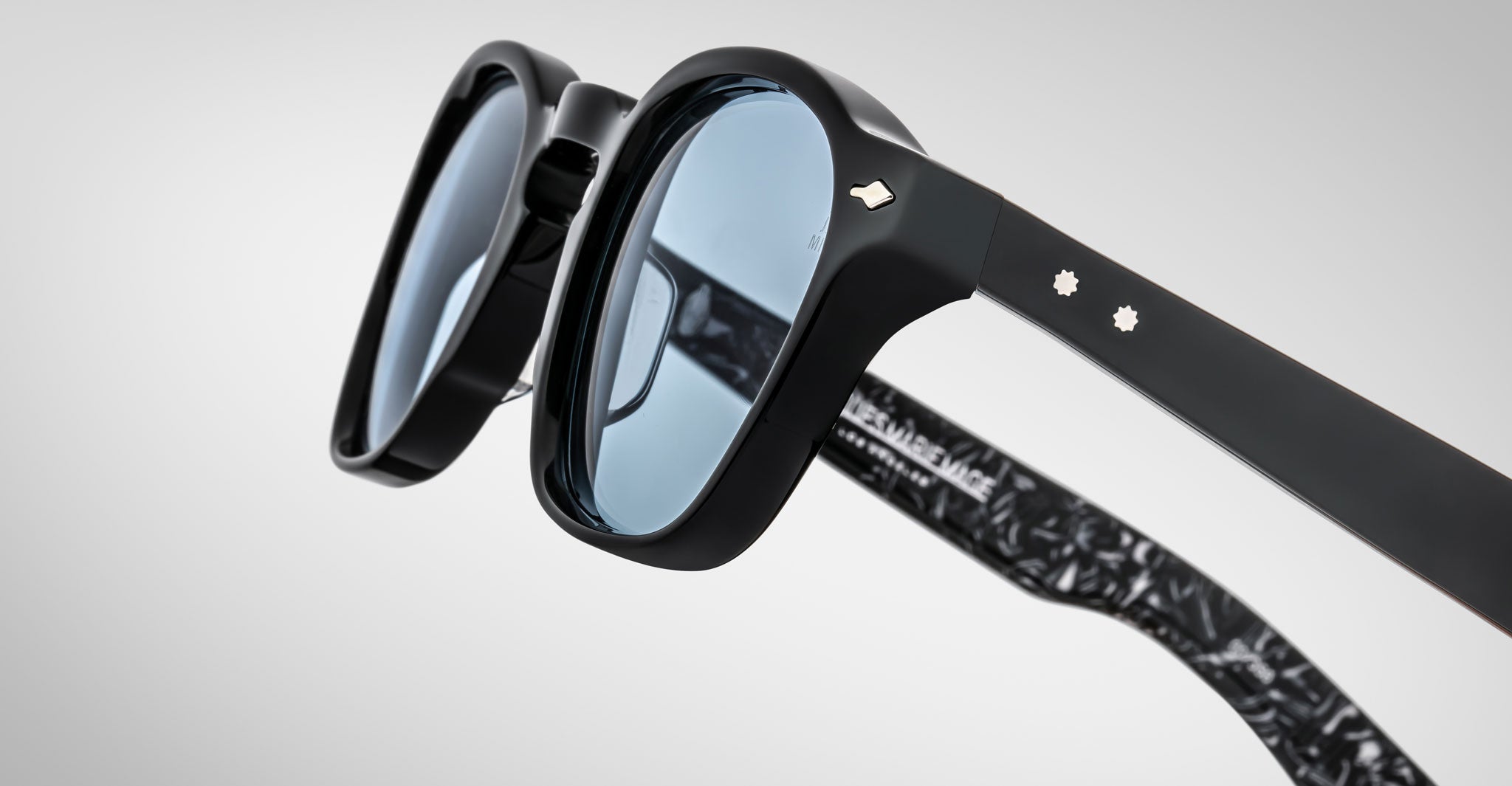 Close-up of Jacques Marie Mage Zephirin sunglasses—limited edition handcrafted acetate eyewear in black with blue tinted lenses, metal rivets on the arms, and a marbled black inner temple pattern.