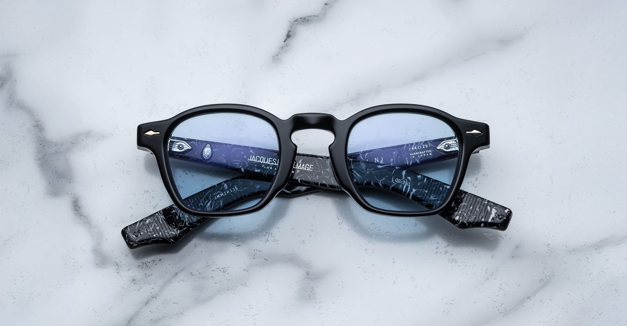 A pair of Jacques Marie Mage Zephirin black-framed eyeglasses with blue-tinted lenses rests on white marble, highlighting the refined craftsmanship of Jacques Marie Mage.