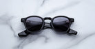 The Jacques Marie Mage Zephirin sunglasses, limited edition with black frames and dark lenses, are showcased on a white marble surface.