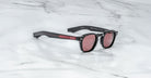 A pair of Jacques Marie Mage Zephirin limited edition black sunglasses with red-tinted lenses and red accents on the arms, inspired by Jacques Marie Mage, displayed on a white marble surface.