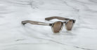 A pair of Jacques Marie Mage Zephirin sunglasses with brown-tinted lenses and transparent gray frames rests on white marble, highlighting this limited edition Jacques Marie Mage eyewear.