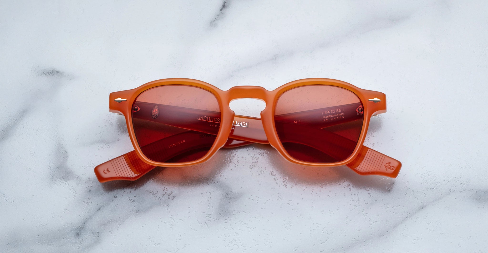 Jacques Marie Mage Zephirin limited edition eyewear, featuring orange frames and red-tinted lenses, is displayed on white marble, showcasing the bold craftsmanship of Jacques Marie Mage.