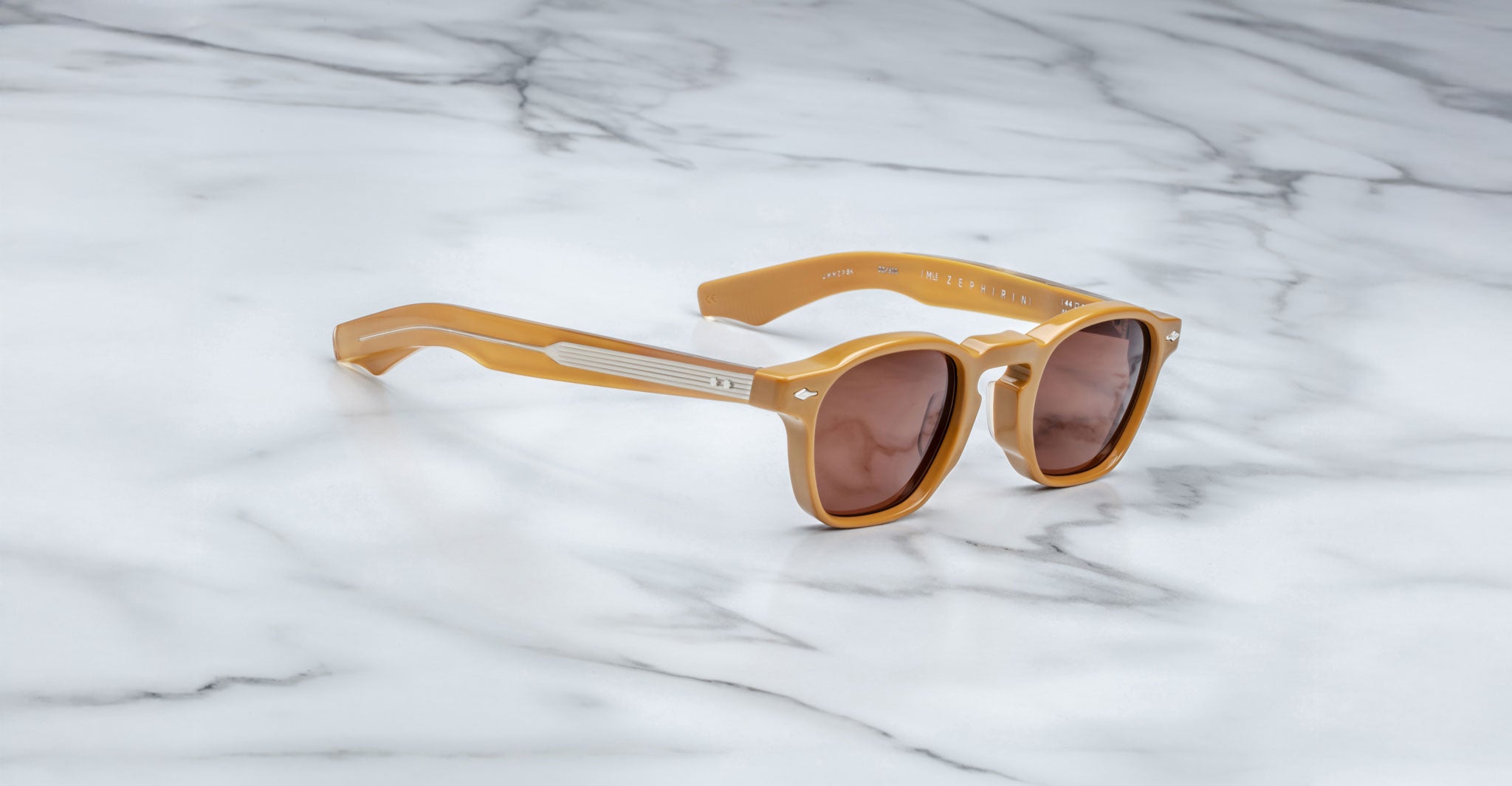 A pair of Jacques Marie Mage Zephirin sunglasses with tan frames and brown lenses rests on a white marble surface.