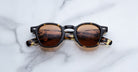 Jacques Marie Mage Zephirin sunglasses—limited edition round tortoiseshell frames with brown lenses handcrafted from acetate—are elegantly displayed on a white marble surface.