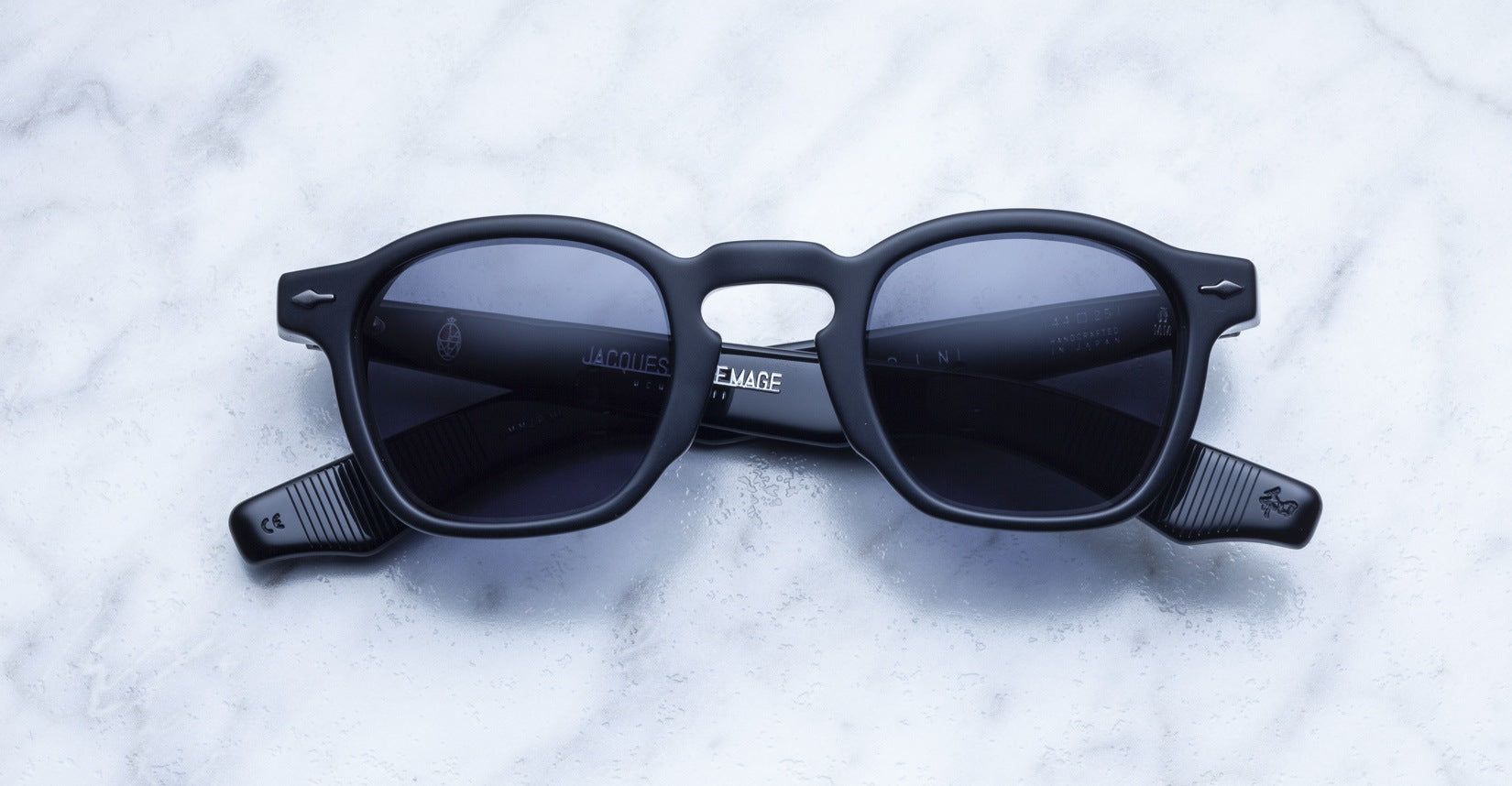 The Jacques Marie Mage Zephirin sunglasses by Jacques Marie Mage feature black handcrafted acetate frames and dark lenses, showcased on white marble. This limited edition eyewear epitomizes unique sophistication.