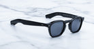 A pair of Jacques Marie Mage Zephirin black sunglasses with dark lenses and handcrafted acetate frames sits on a white marble surface.