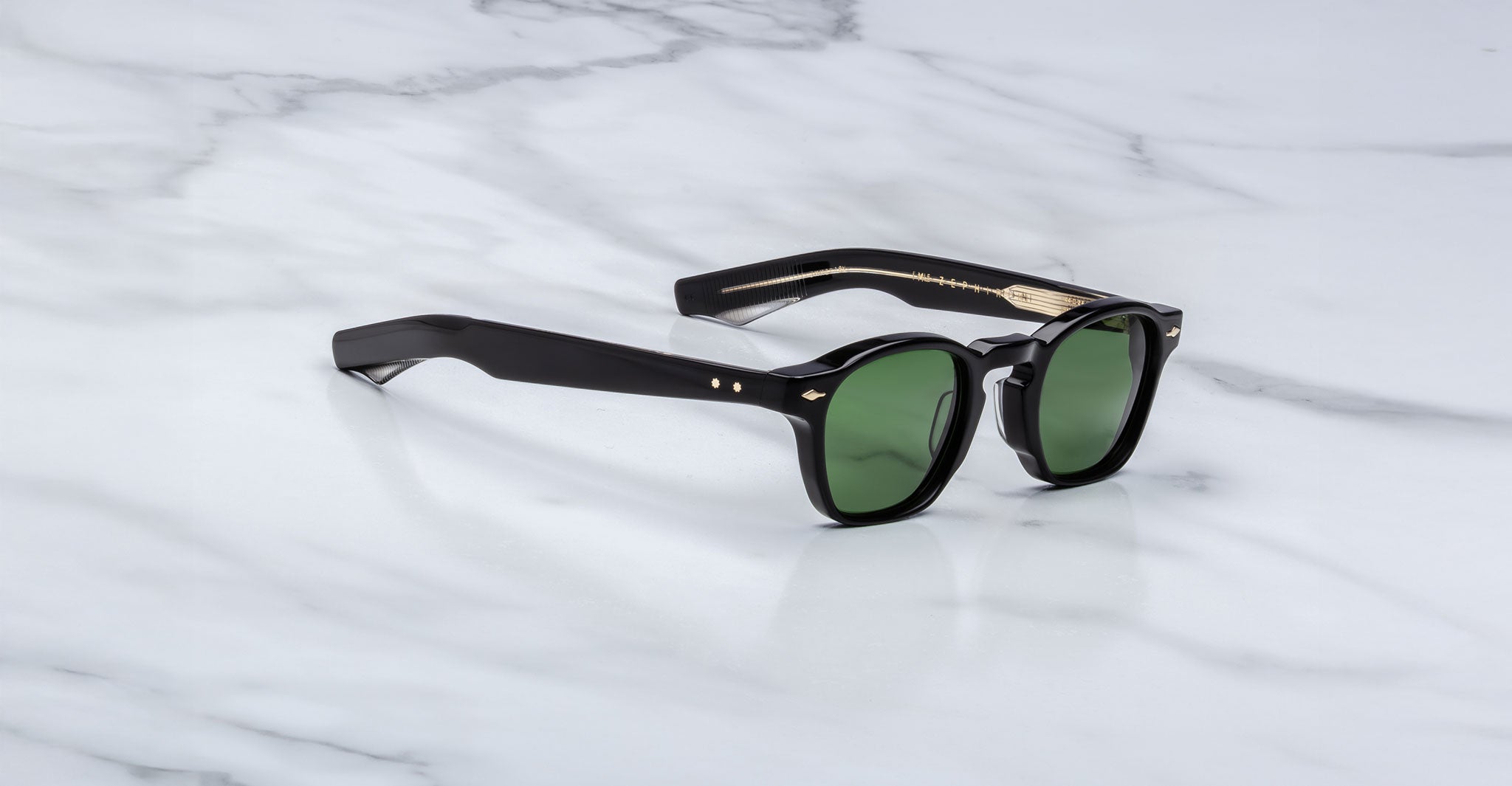 The Jacques Marie Mage Zephirin sunglasses feature green lenses and gold-detailed arms, showcased on a white marble surface, reflecting the elegance of limited edition Jacques Marie Mage handcrafted acetate eyewear.