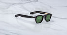 The Jacques Marie Mage Zephirin sunglasses feature green lenses and gold-detailed arms, showcased on a white marble surface, reflecting the elegance of limited edition Jacques Marie Mage handcrafted acetate eyewear.