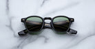 The Jacques Marie Mage Zephirin sunglasses by Jacques Marie Mage, featuring black round frames and green lenses reminiscent of handcrafted acetate, are displayed on a white marble surface.