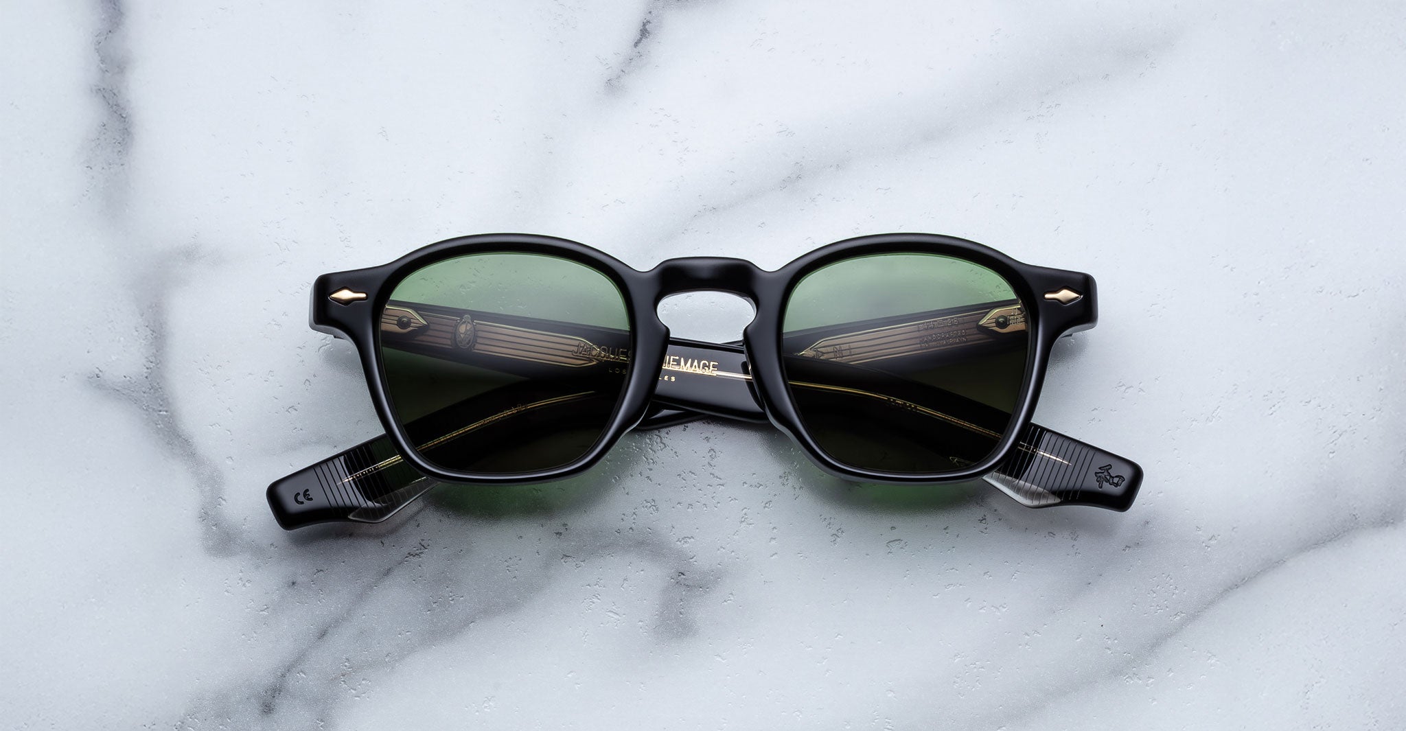 The Jacques Marie Mage Zephirin sunglasses by Jacques Marie Mage, featuring black round frames and green lenses reminiscent of handcrafted acetate, are displayed on a white marble surface.