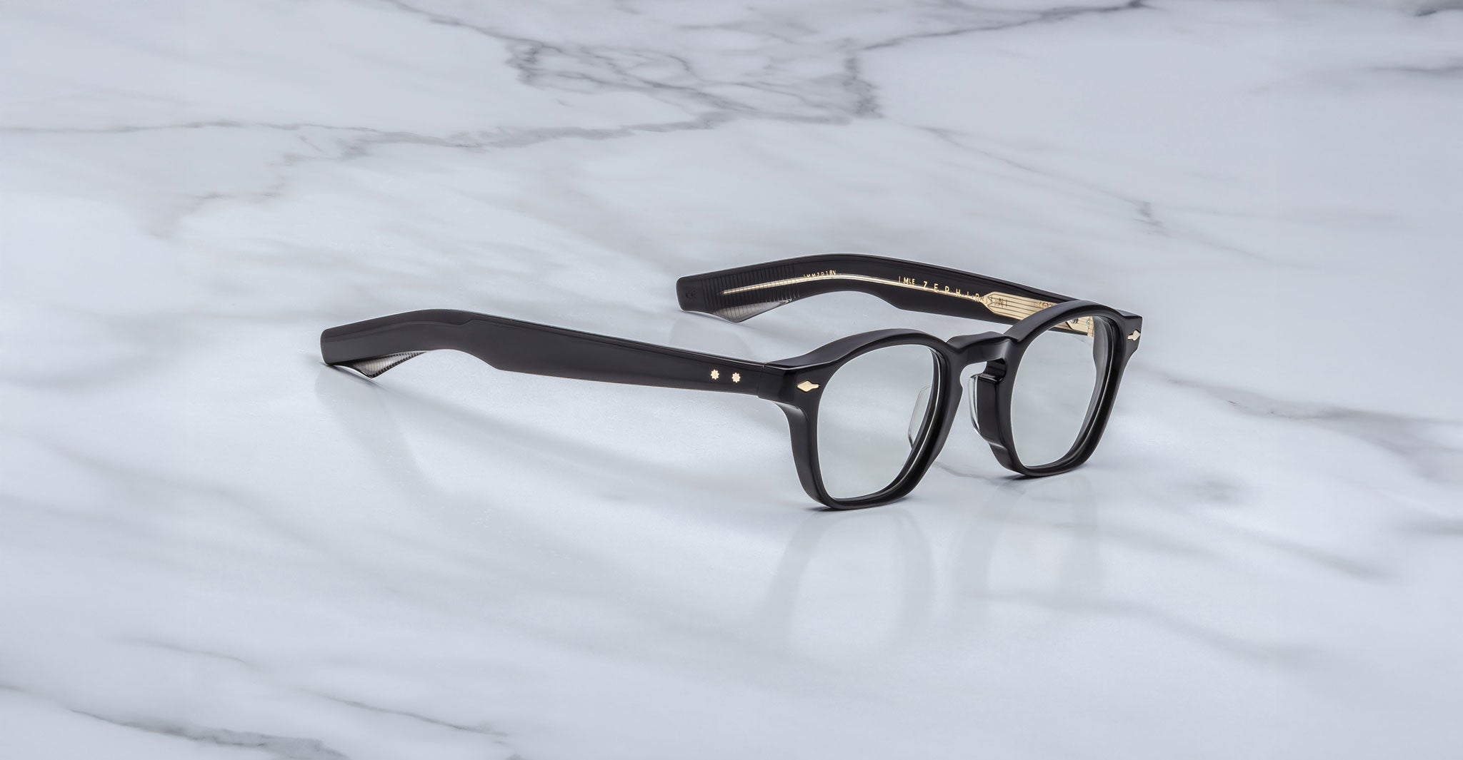 Jacques Marie Mage Zephirin black-framed handcrafted acetate spectacles with clear lenses and gold accents by Jacques Marie Mage, displayed on a white marble surface.