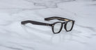 Jacques Marie Mage Zephirin black-framed handcrafted acetate spectacles with clear lenses and gold accents by Jacques Marie Mage, displayed on a white marble surface.