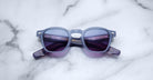 A pair of Jacques Marie Mage Zephirin sunglasses featuring square blue frames and purple-tinted lenses rests on a white marble surface, highlighting the brand’s exquisite handcrafted acetate eyewear.