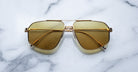 The Jacques Marie Mage Jagger sunglasses by Jacques Marie Mage, with oversized gold beta titanium frames and yellow-tinted lenses, are displayed on a white marble surface.