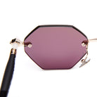A close-up of a stylish sunglass lens with a geometric, hexagonal shape, purple tint, and gold metal frame with a black temple and white detail near the hinge.