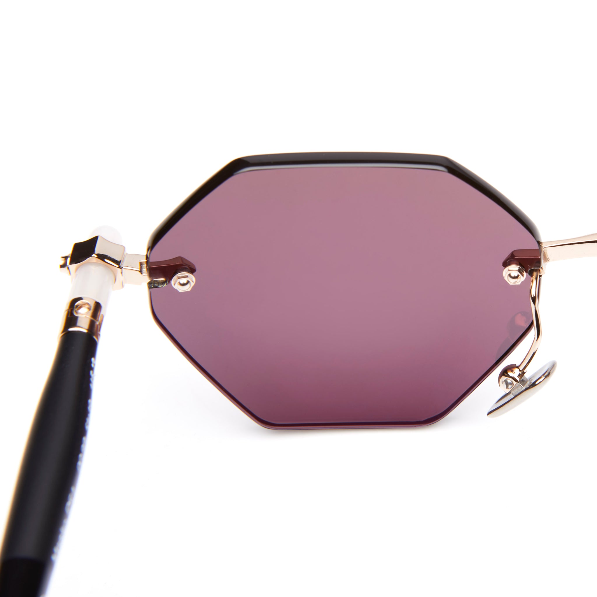 A close-up of a stylish sunglass lens with a geometric, hexagonal shape, purple tint, and gold metal frame with a black temple and white detail near the hinge.